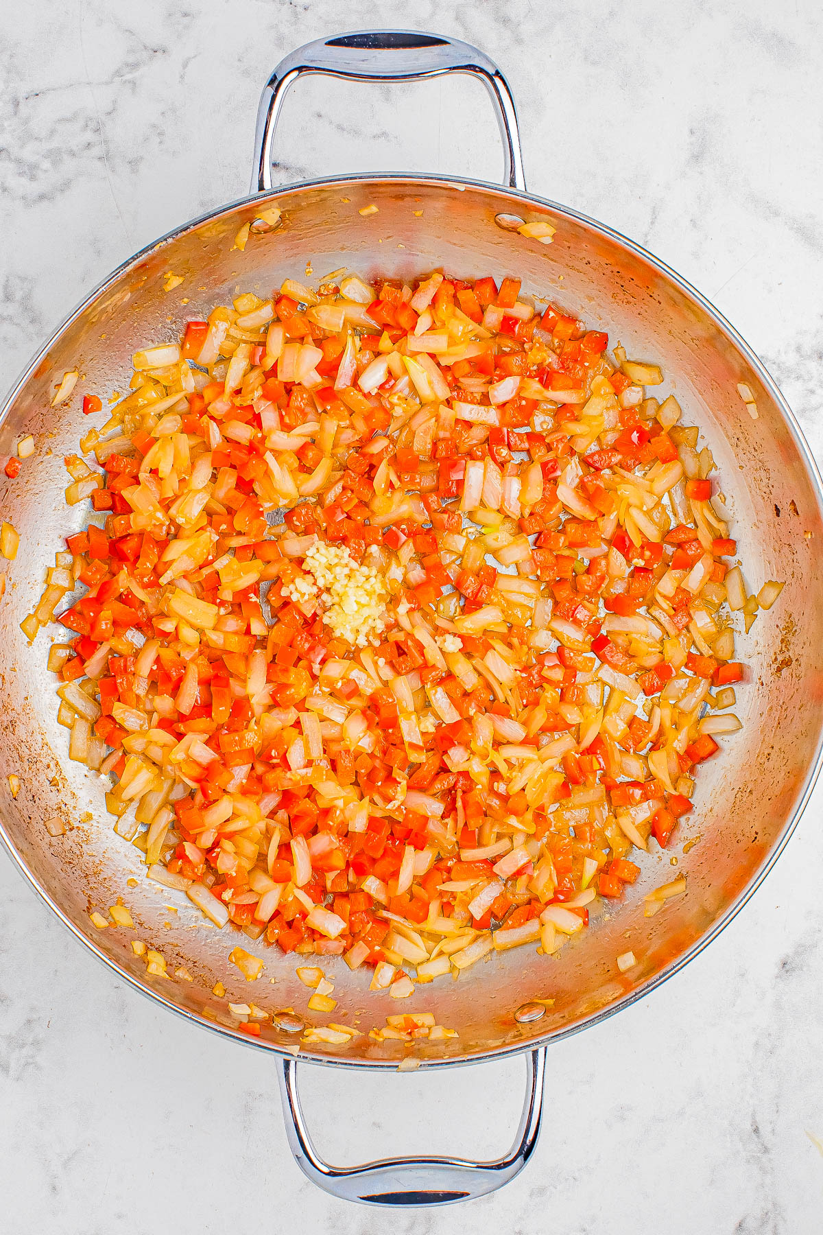 A skillet filled with sautéed diced onions, red bell peppers, and minced garlic on a marble countertop.