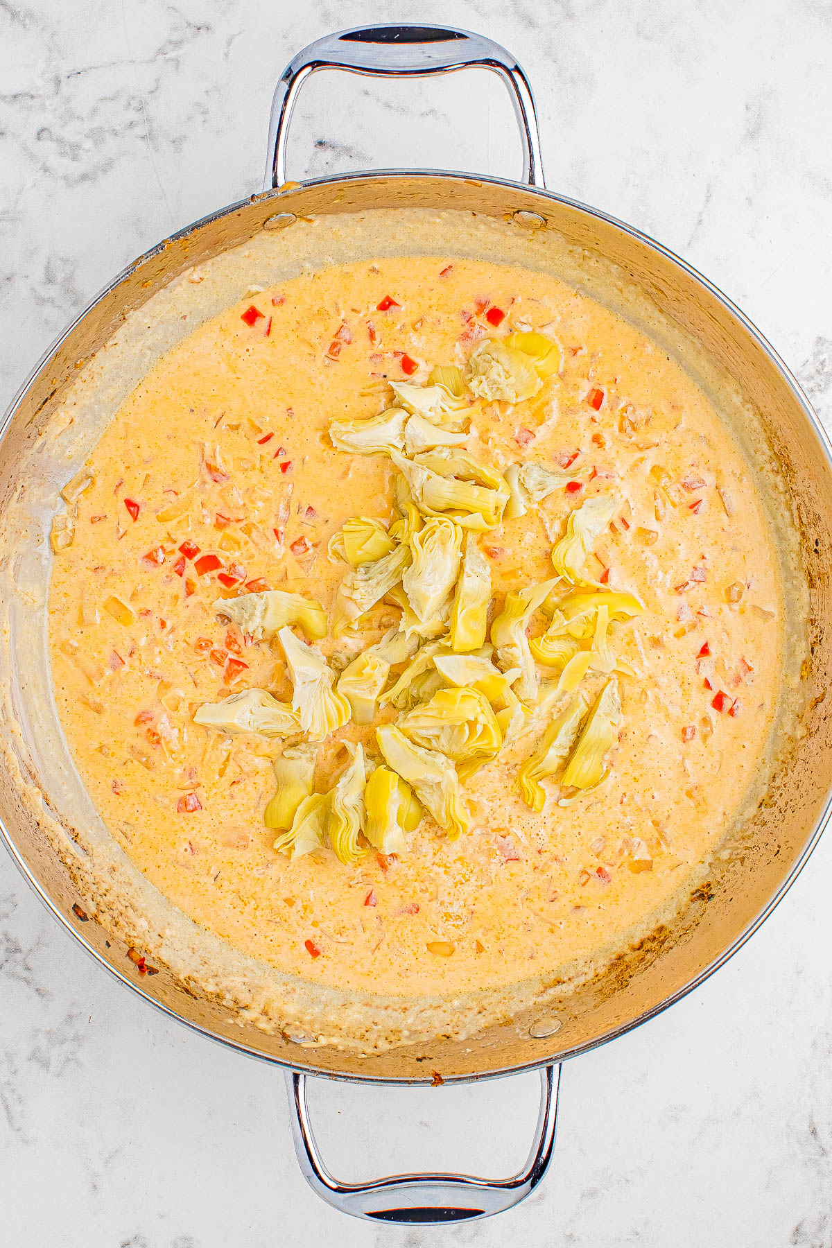 A metal skillet containing a creamy sauce with visible chunks of artichoke hearts, red bell peppers, and other ingredients on a marble countertop.