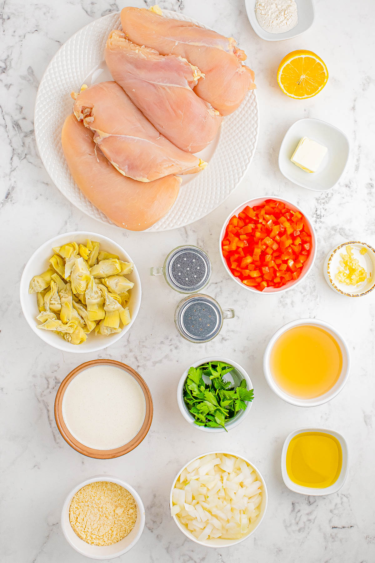 Top-down view of various ingredients on a white marble surface, including raw chicken breasts, chopped vegetables, seasonings, and small bowls containing liquids, lemon, and butter.