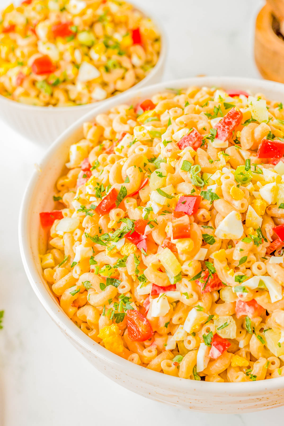 A close-up view of a bowl filled with macaroni salad mixed with diced tomatoes, bell peppers, hard-boiled eggs, and fresh herbs. Another similar bowl is partly visible in the background.