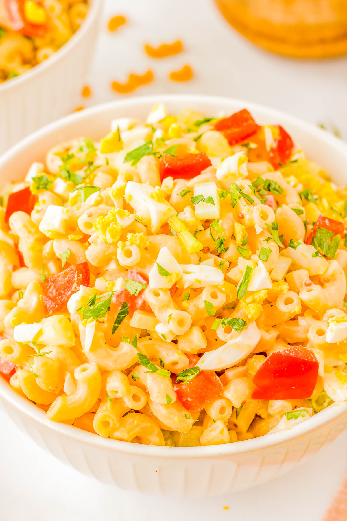 A bowl filled with macaroni salad, topped with diced red bell peppers, chopped celery, hard-boiled eggs, and garnished with fresh herbs.