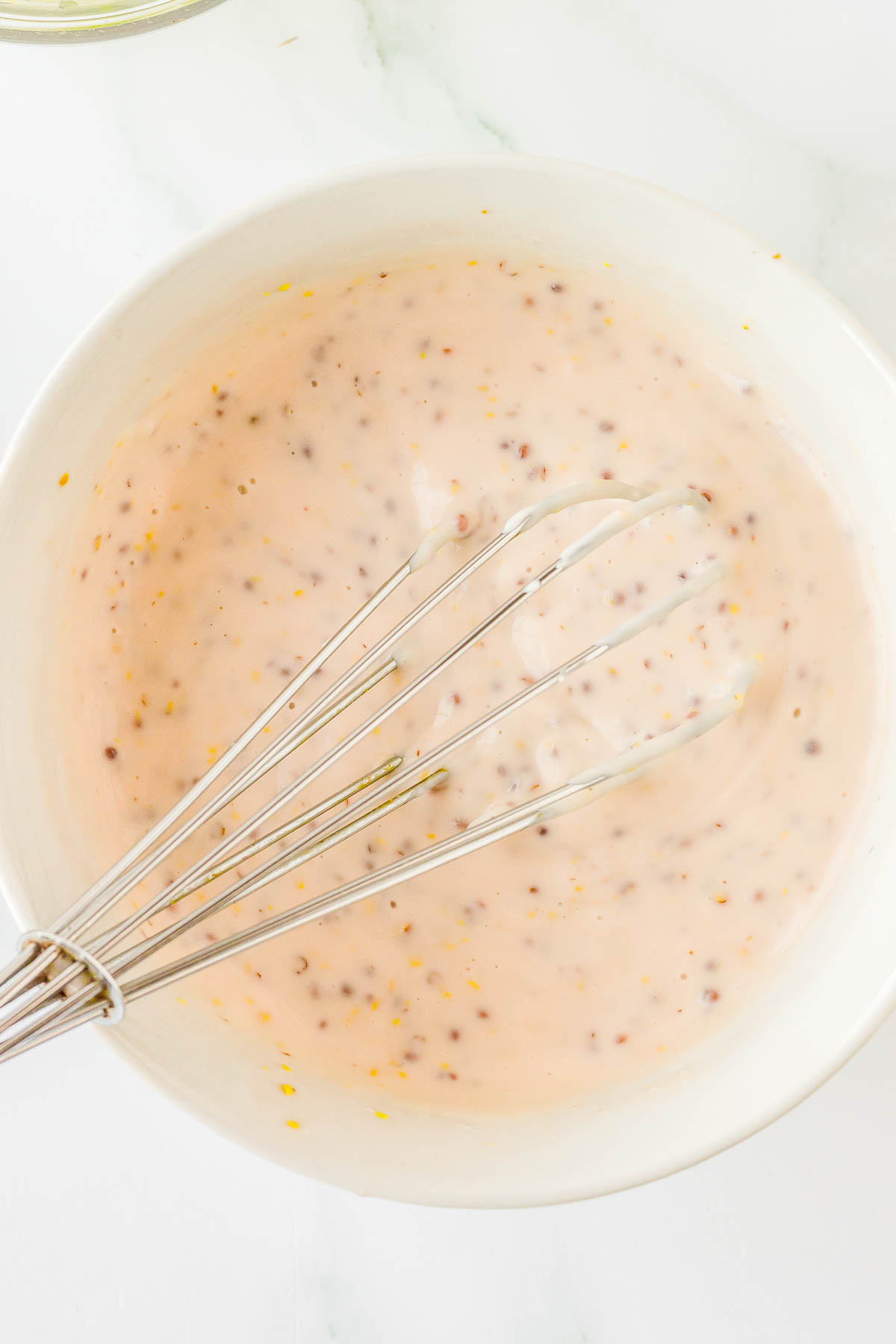 Whisk resting in a white bowl containing a creamy, speckled dressing or sauce.