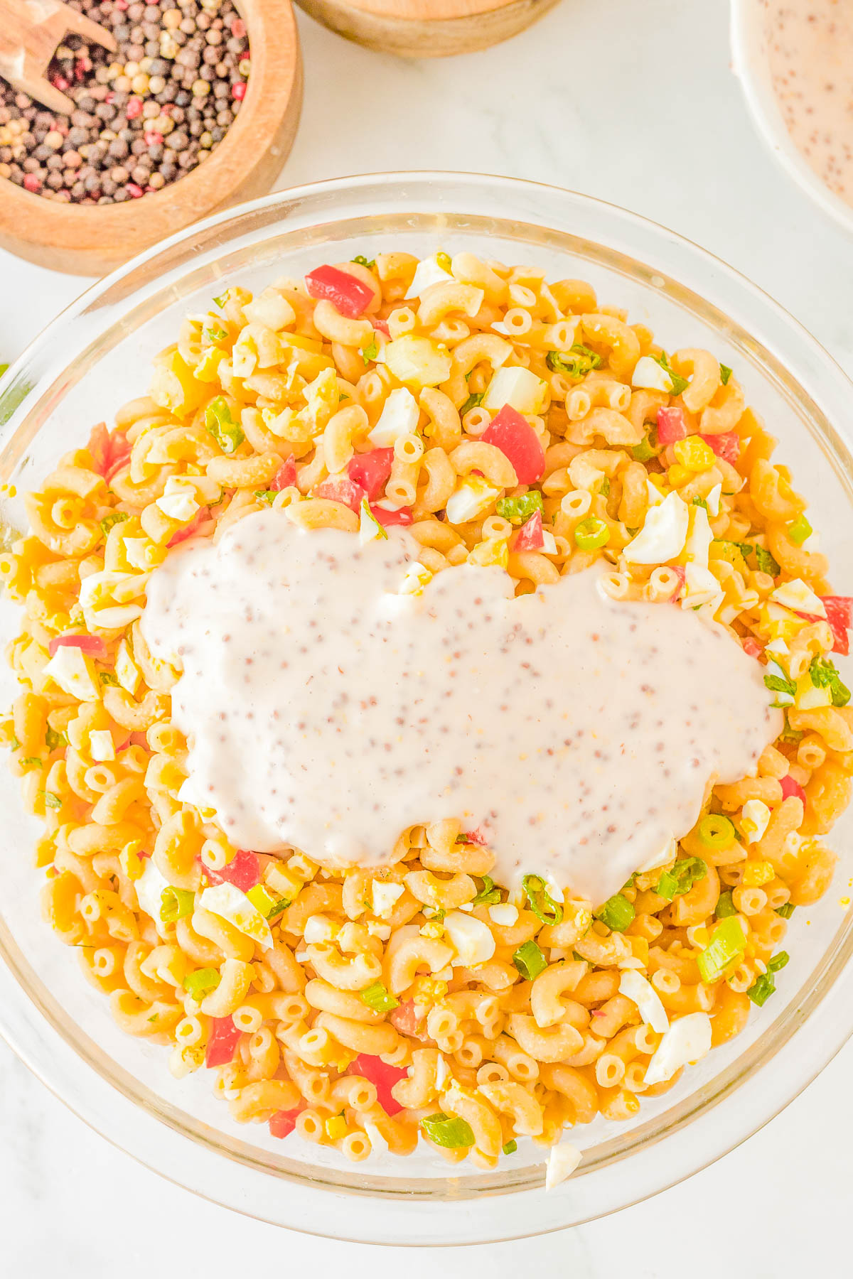 A bowl of macaroni salad with diced vegetables and a creamy dressing on top, placed on a white surface next to a small bowl of peppercorns.