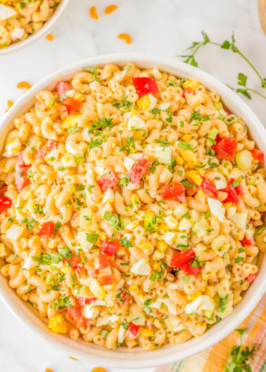 A bowl of macaroni salad with diced tomatoes, yellow bell peppers, chopped eggs, and herbs on a white surface.