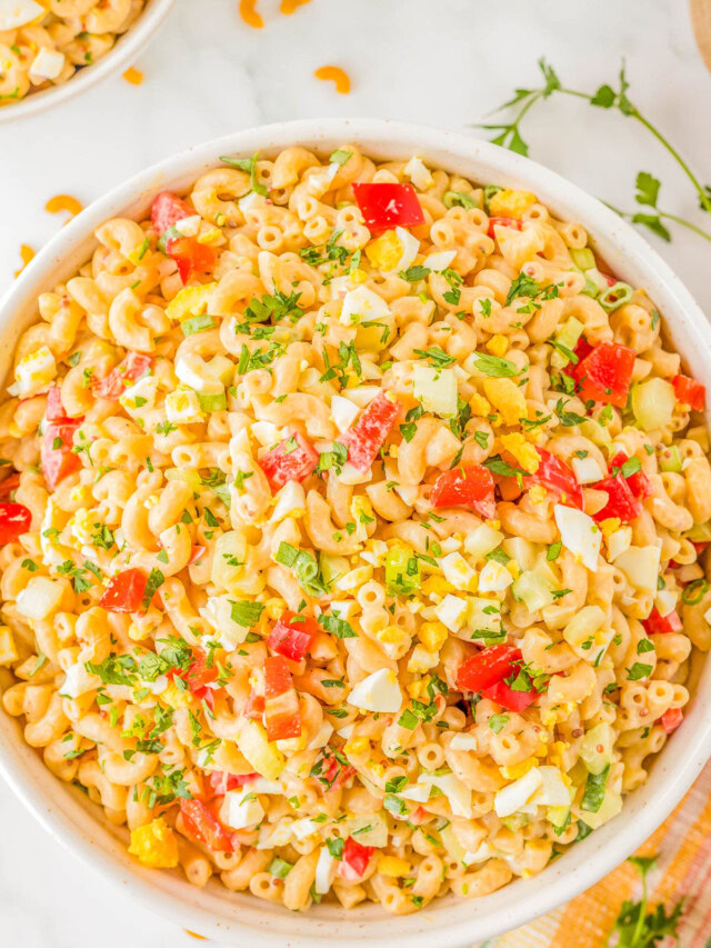 A bowl of macaroni salad with diced tomatoes, yellow bell peppers, chopped eggs, and herbs on a white surface.