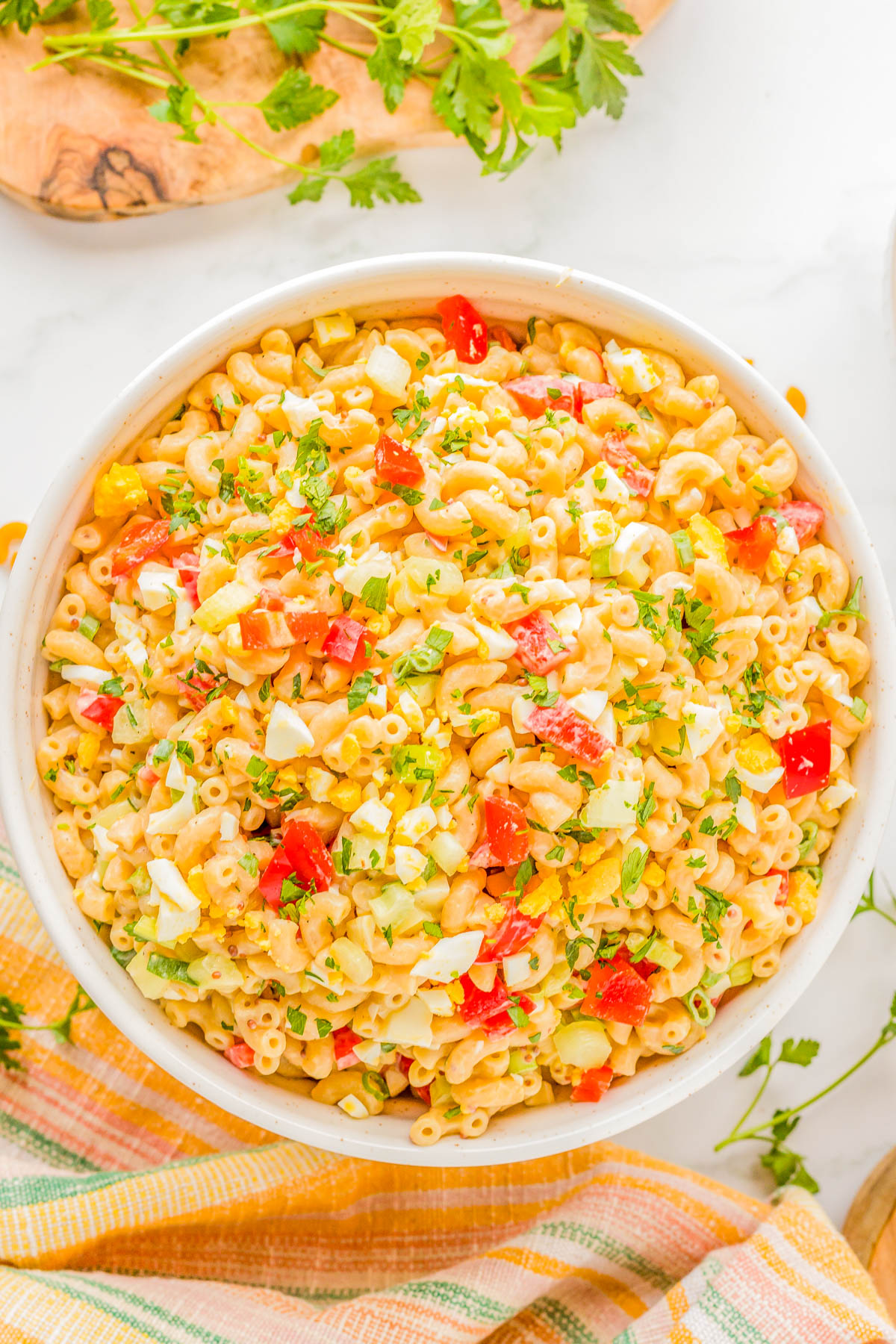 A large bowl filled with macaroni salad featuring chopped vegetables and herbs, garnished with parsley, placed on a colorful striped towel with a wooden cutting board and fresh parsley in the background.