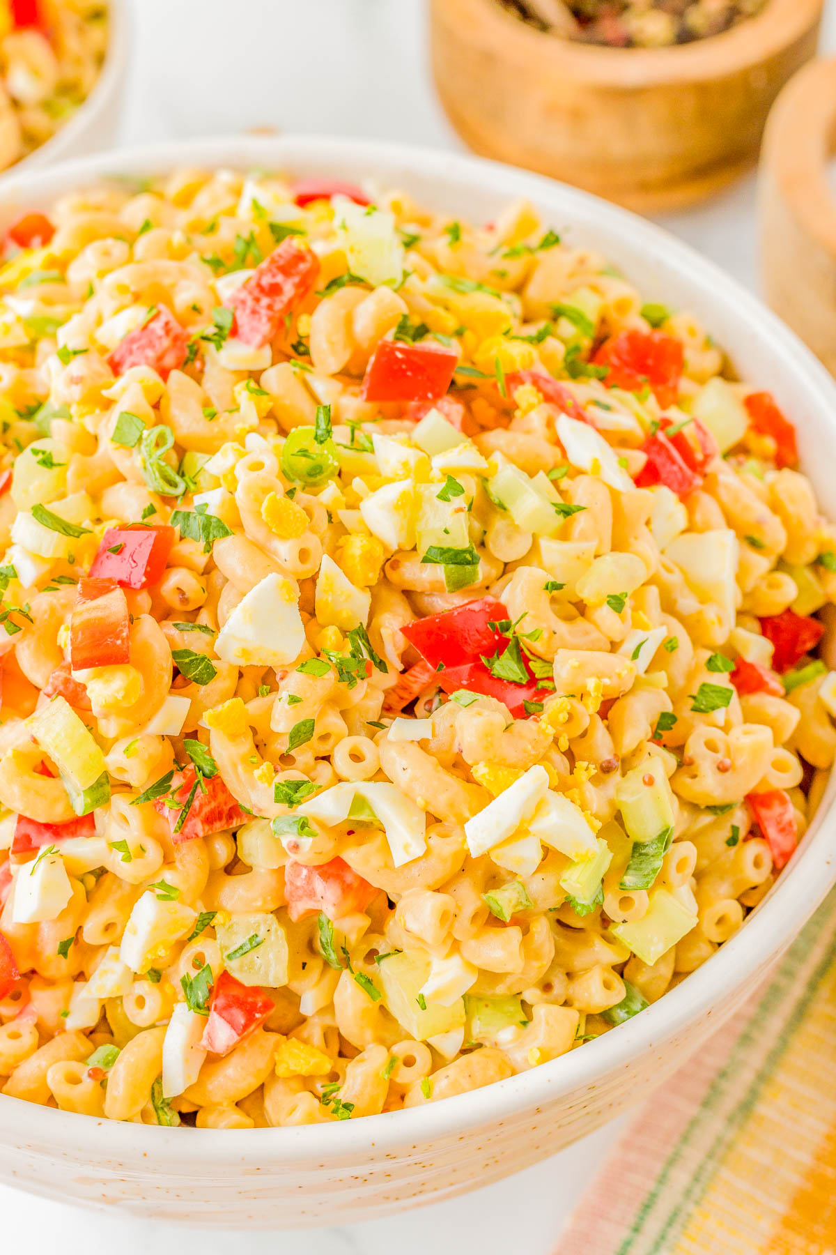 A bowl of macaroni salad with diced red bell peppers, chopped hard-boiled eggs, celery, and parsley, set on a white surface with additional bowls in the background.