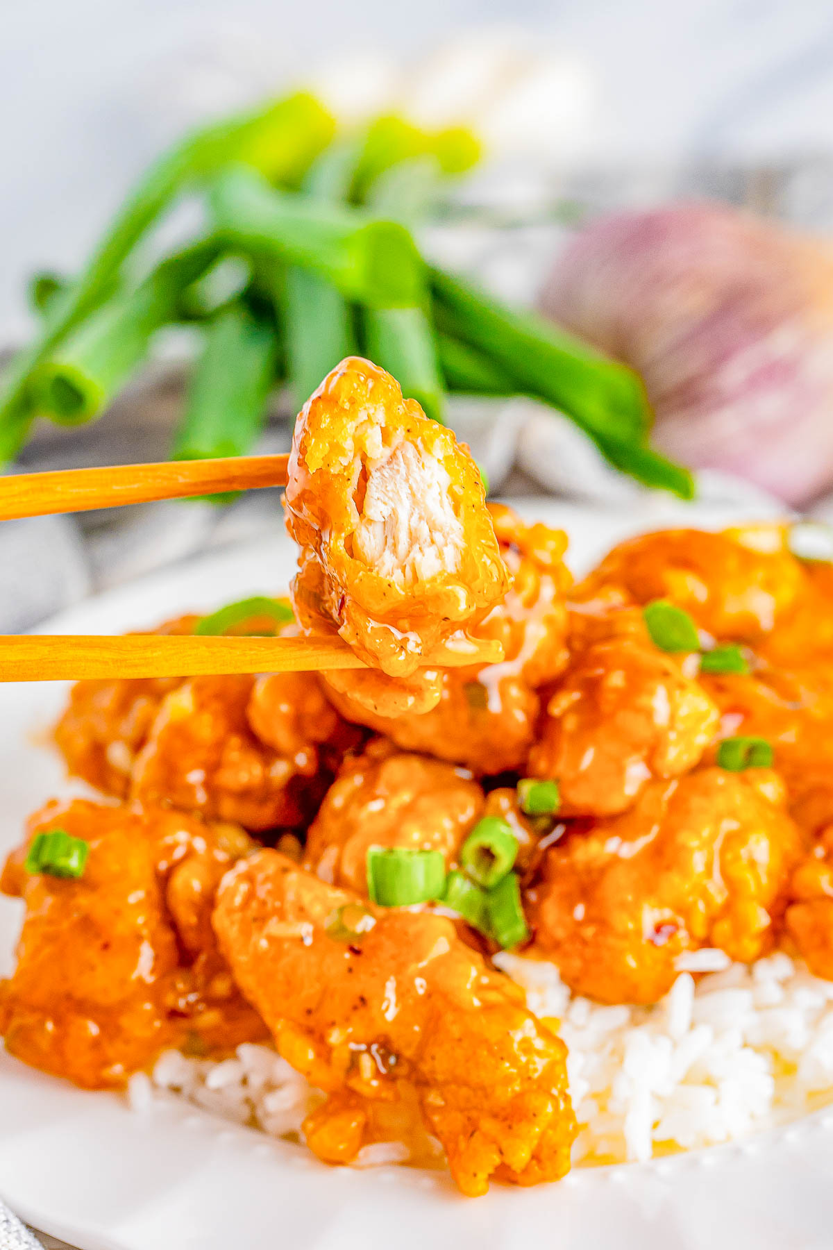 A plate of fried chicken pieces coated in orange sauce, served on white rice. A piece of chicken is held by chopsticks. Green onions and sliced garlic are visible in the background.