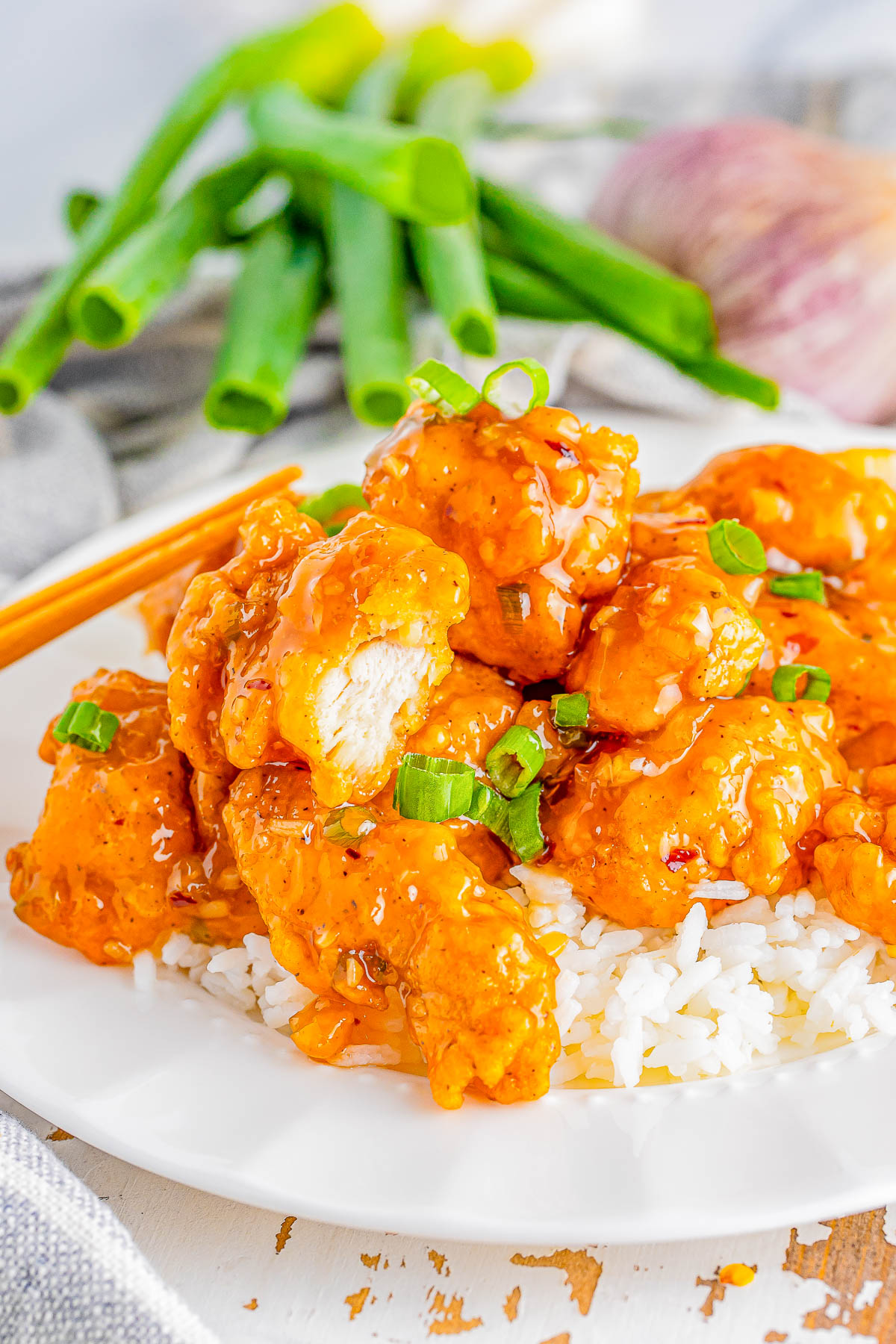 A plate of white rice topped with crispy, glazed chicken pieces garnished with chopped green onions, with green onions and an onion in the background.