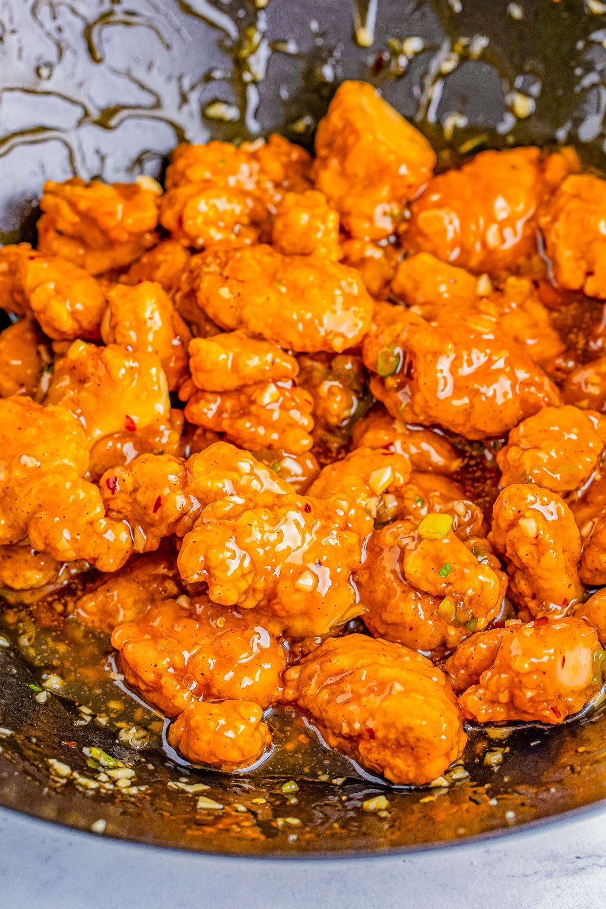 Close-up of a pan filled with battered and fried chicken pieces coated in a sticky orange sauce, topped with finely chopped green onions and sesame seeds.