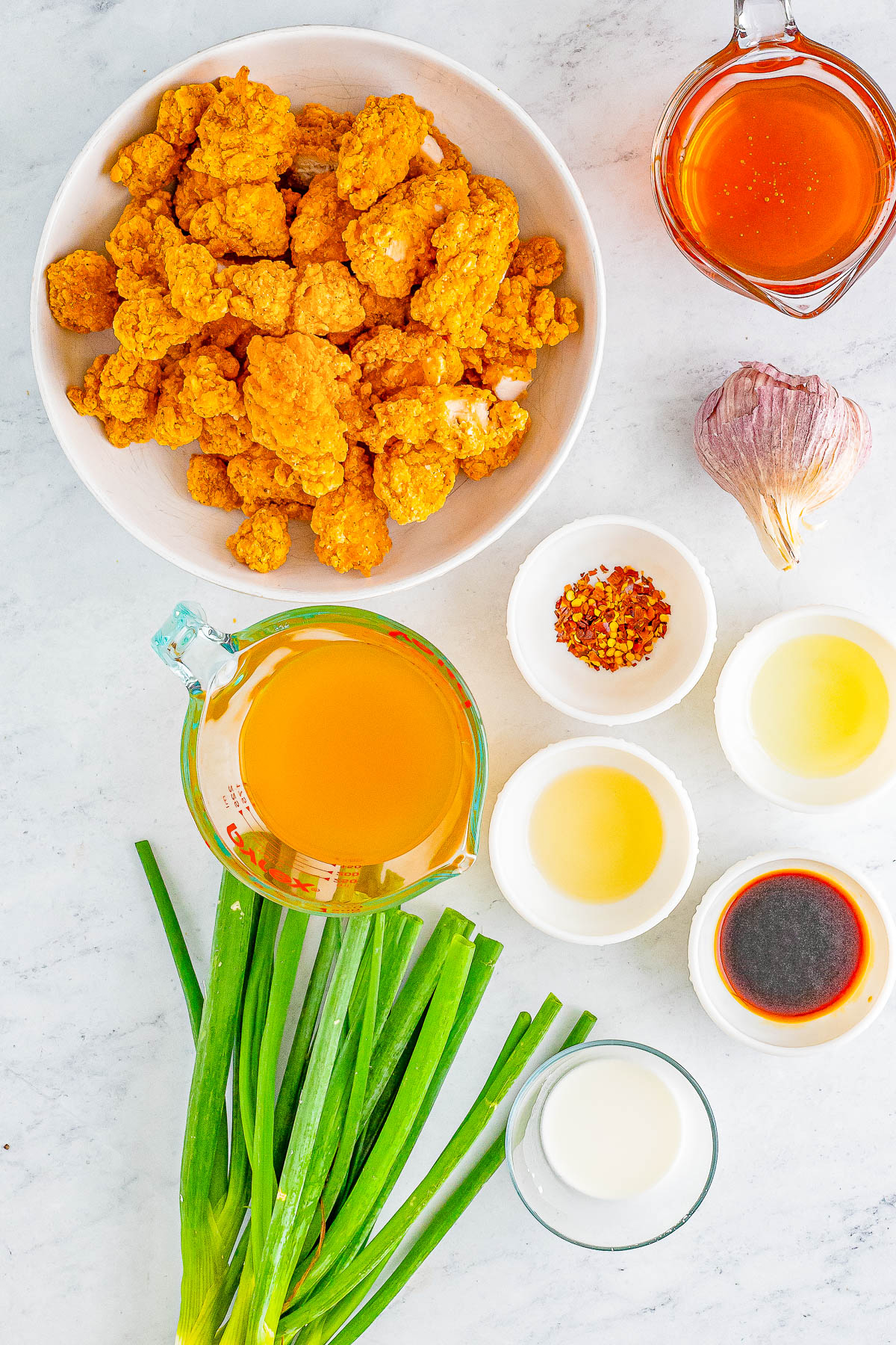 An arrangement of ingredients including fried chicken pieces, garlic, spring onions, and various liquids in measuring cups and bowls on a white marble surface.