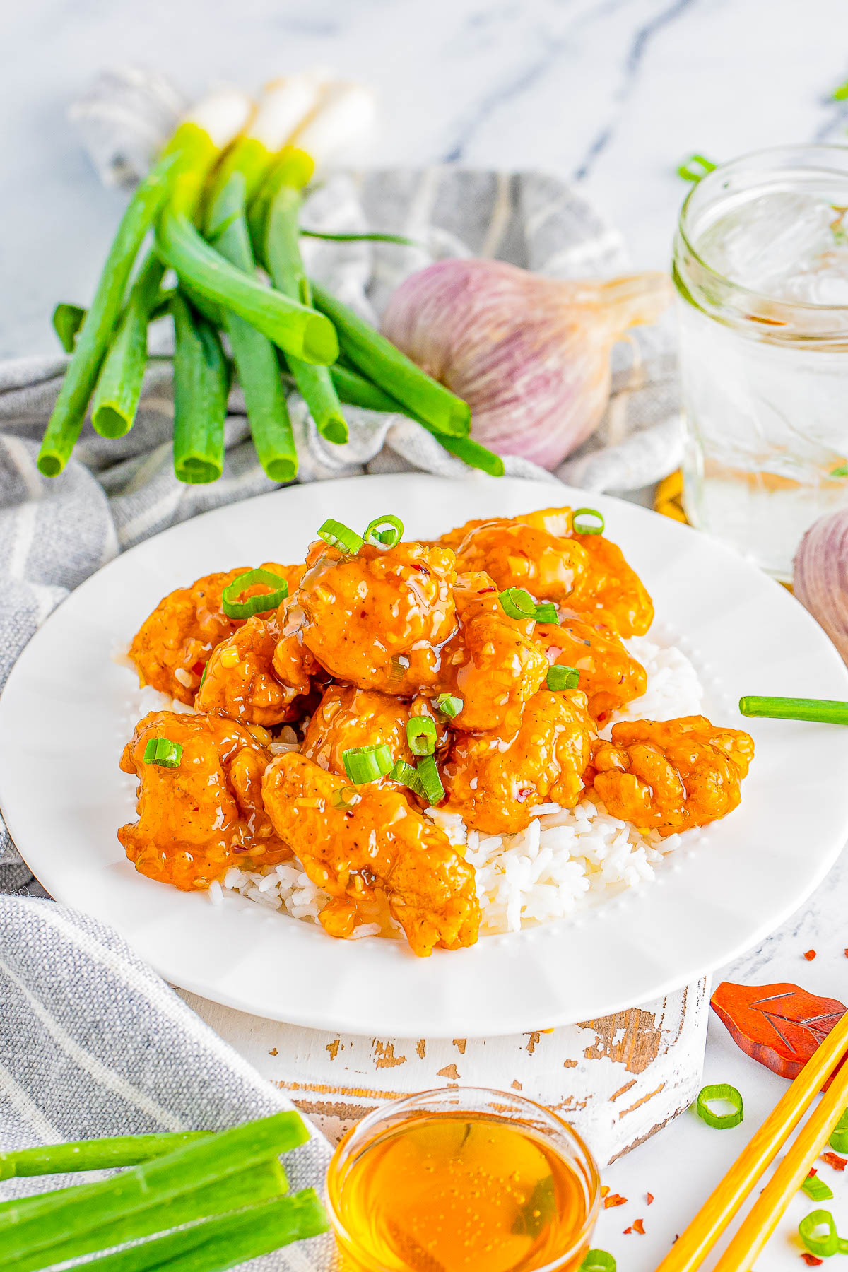 A white plate with orange-colored crispy chicken pieces garnished with green onions placed on a bed of white rice. Fresh green onions, garlic, a glass of water, and a small bowl of sauce are nearby.