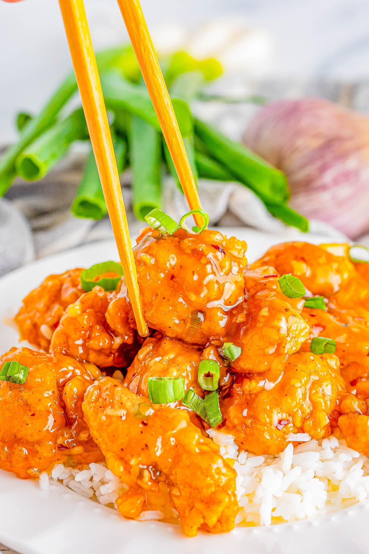 A pair of chopsticks lifts a piece of sauced chicken garnished with chopped green onions from a plate of white rice. Green onions and a red onion are blurred in the background.