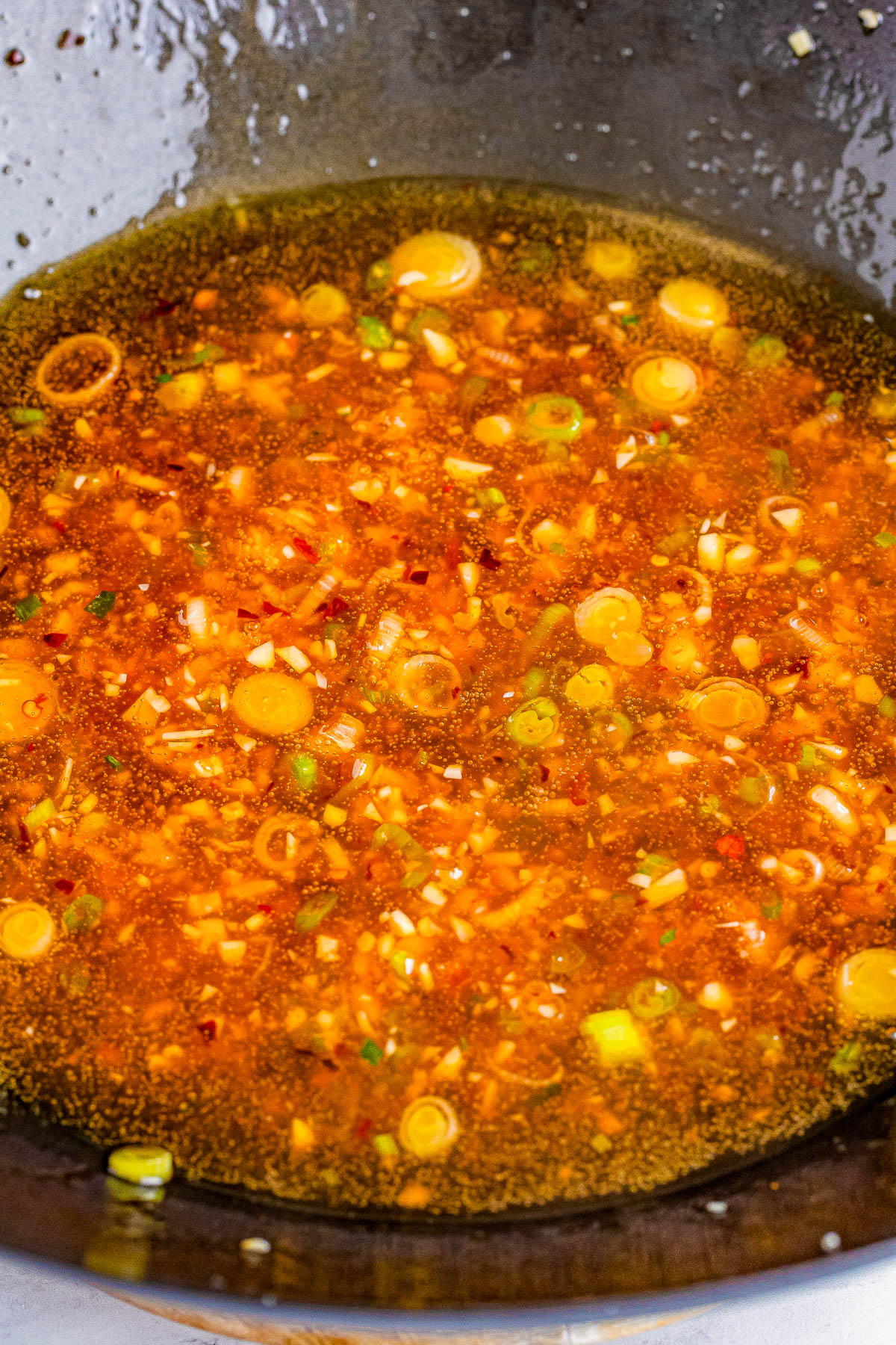 A close-up of a simmering mixture of minced garlic, ginger, chopped green onions, and red chili flakes in a hot wok.