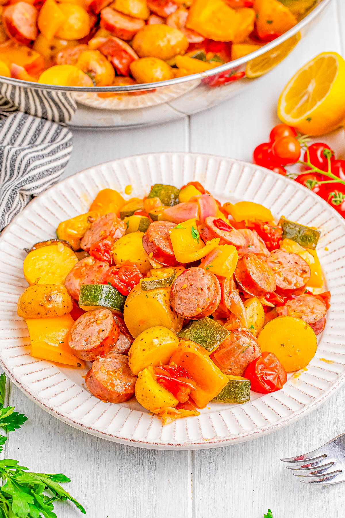 A plate of cooked sausages, potatoes, bell peppers, zucchini, and cherry tomatoes in a seasoned sauce. A skillet with more of the dish is in the background, along with a halved lemon and herbs.