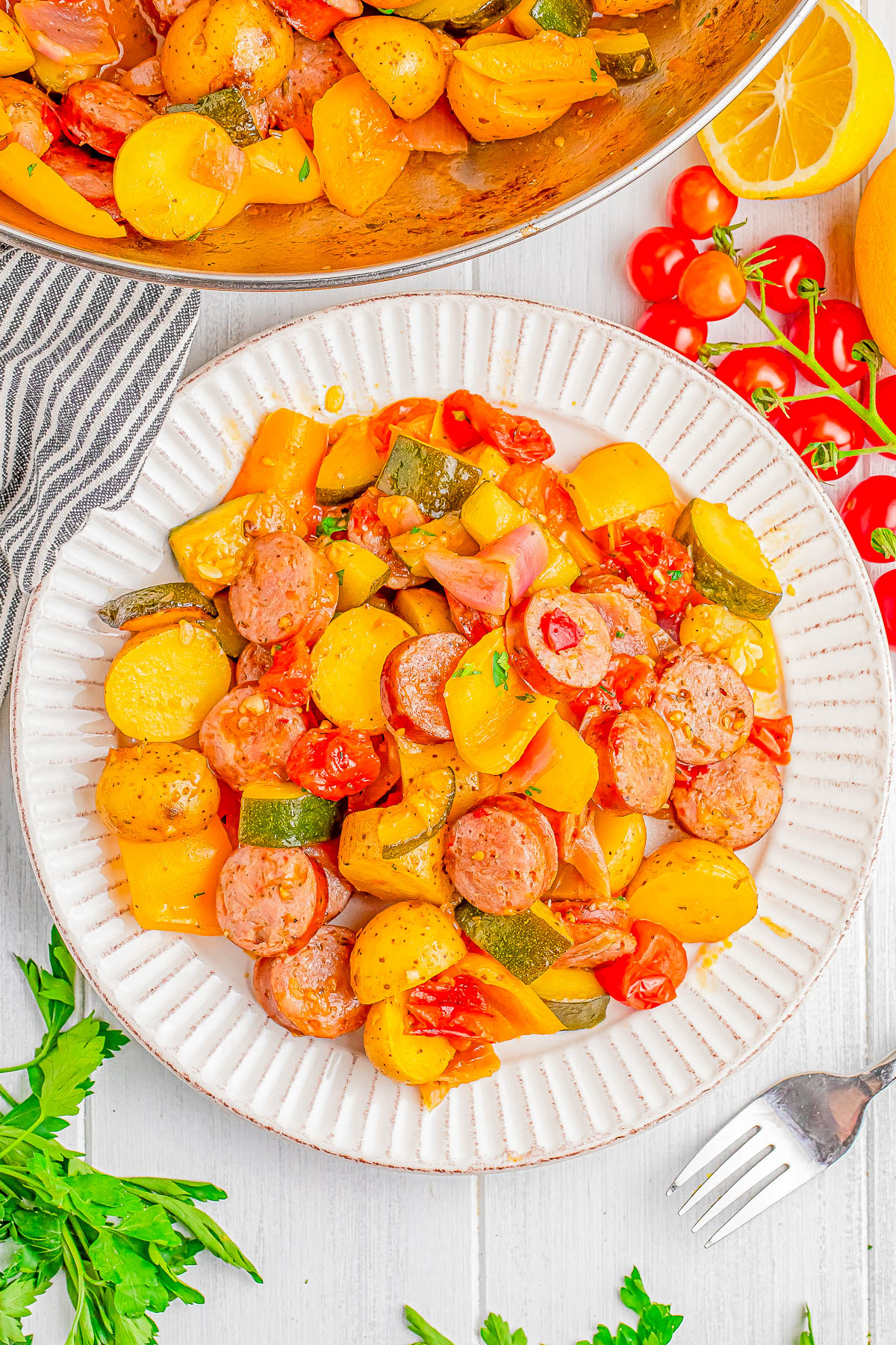 A white plate contains a dish of sliced sausages, yellow potatoes, zucchini, onions, and tomatoes. A fork is on the side, with additional ingredients scattered around, including parsley and a lemon.