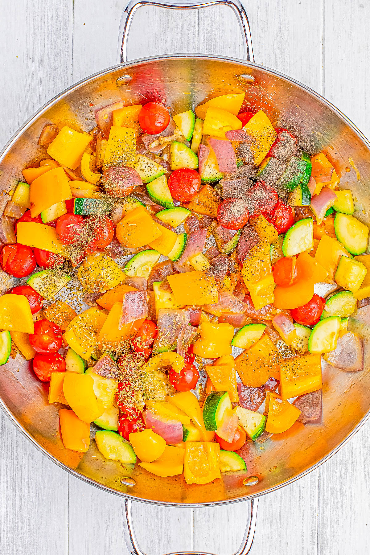 A large pan filled with a colorful mix of diced vegetables including yellow bell peppers, cherry tomatoes, zucchini, and red onions, seasoned with salt, pepper, and dried herbs.