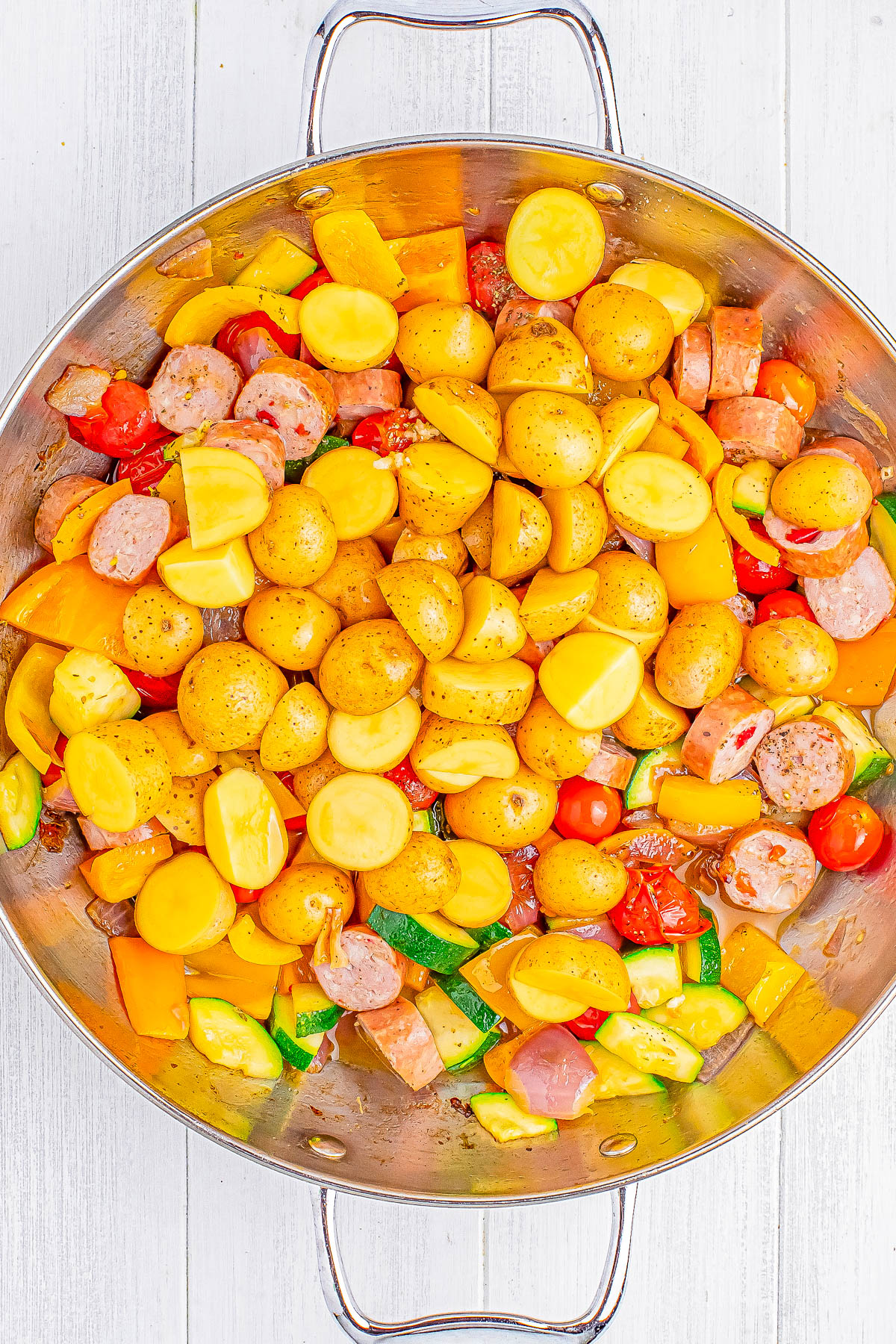 A stainless steel pan containing halved baby potatoes, sliced sausage, and assorted vegetables including bell peppers and cherry tomatoes. The dish appears to be seasoned and ready to cook.