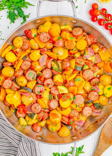 A skillet filled with cooked sausage slices, yellow bell peppers, cherry tomatoes, onions, and diced potatoes, garnished with fresh parsley, with a wooden spoon and herbs on the side.