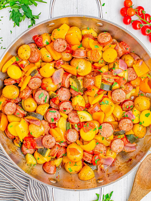 A skillet filled with cooked sausage slices, yellow bell peppers, cherry tomatoes, onions, and diced potatoes, garnished with fresh parsley, with a wooden spoon and herbs on the side.