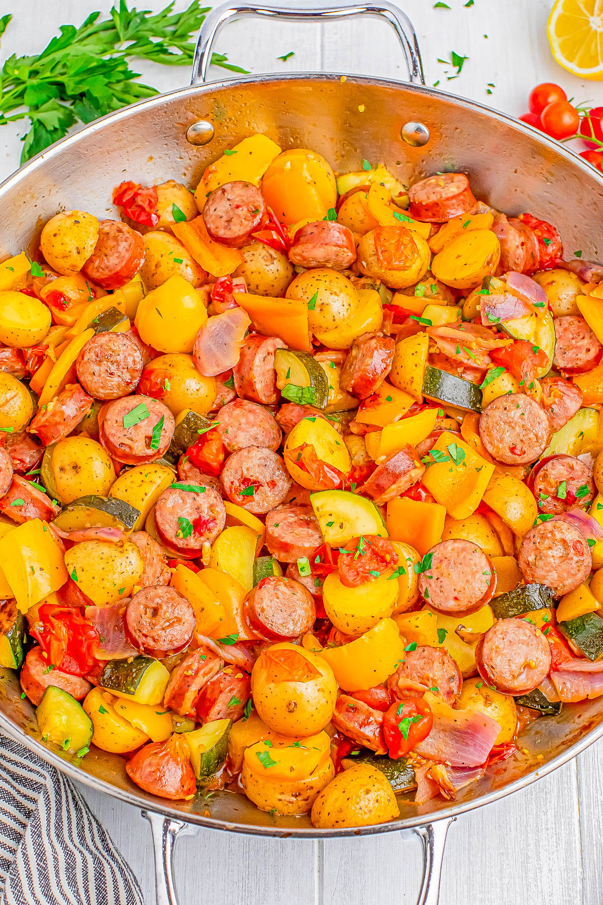 A skillet filled with sliced sausages, diced yellow bell peppers, cherry tomatoes, chopped zucchini, and pieces of potatoes, garnished with chopped parsley.