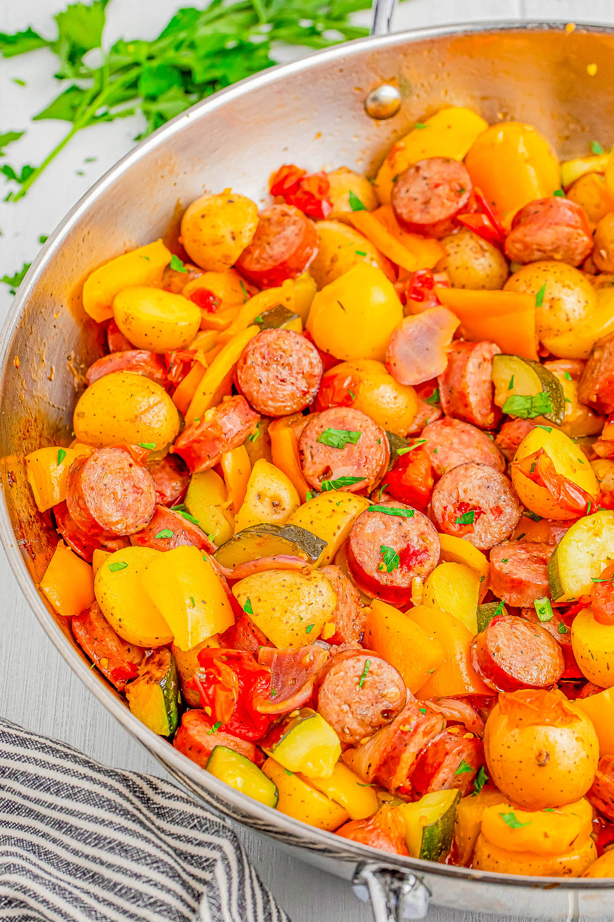 A skillet containing a colorful mix of sliced sausage, yellow bell peppers, baby potatoes, zucchini, and cherry tomatoes, garnished with fresh parsley.