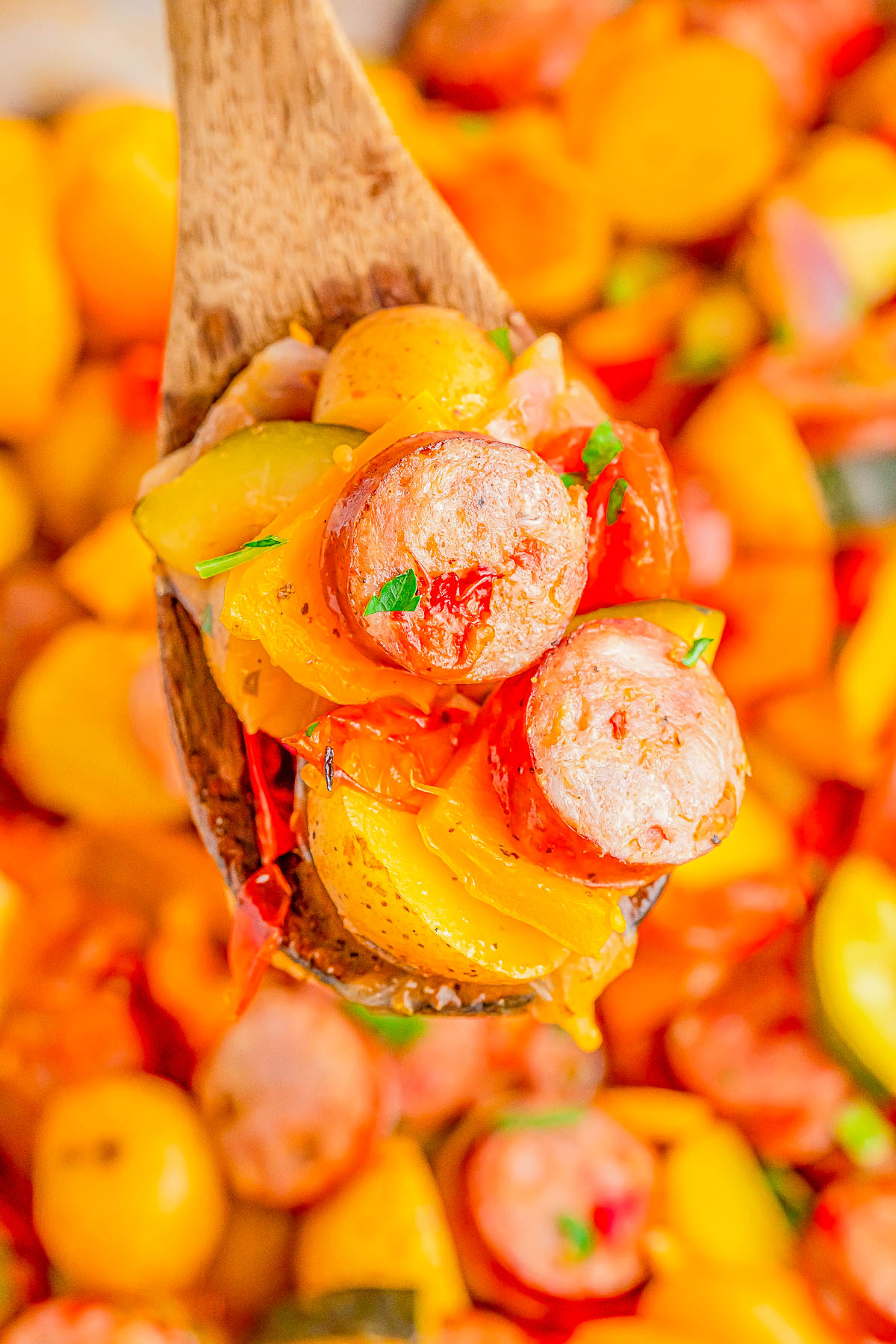 A wooden spoon holds a mixture of roasted sausages, yellow bell peppers, and potatoes garnished with herbs, with more of the dish visible in the background.