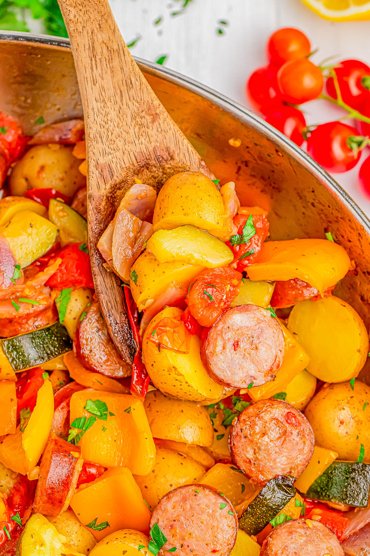 A wooden spoon scooping a dish of roasted vegetables and sausage, including potatoes, zucchini, and bell peppers, garnished with herbs.