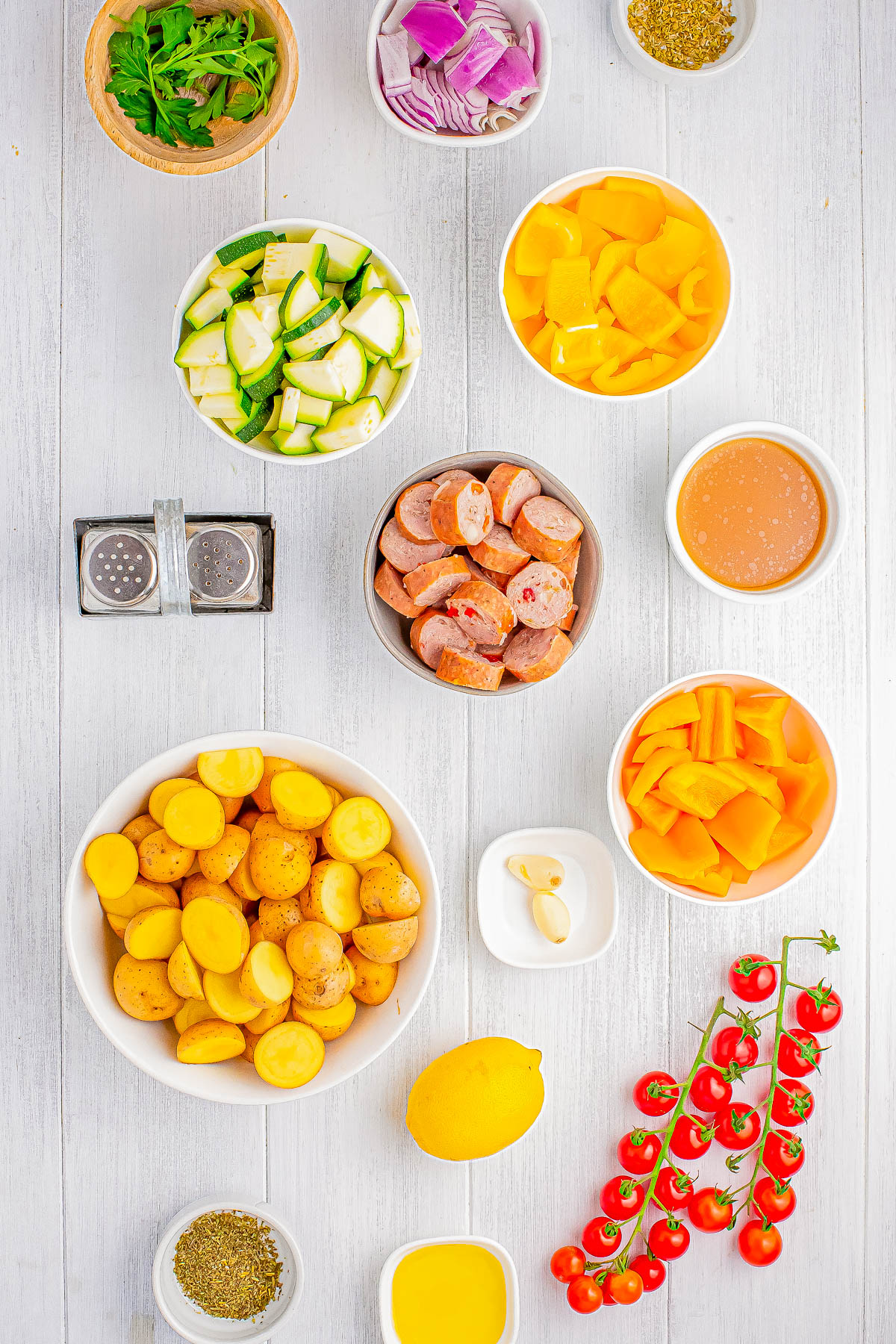 Various ingredients including chopped vegetables, sausage, potatoes, spices, herbs, lemon, cherry tomatoes, and sauces arranged on a white wooden surface.