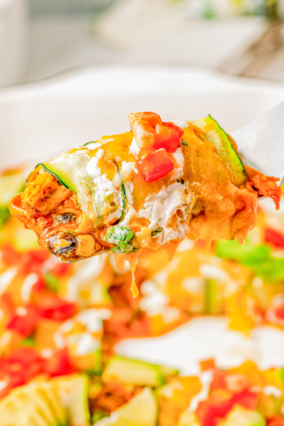 A serving spoon lifting a portion of vegetable lasagna topped with cheese, sour cream, and diced tomatoes. The dish is in a white casserole dish with additional portions visible in the background.