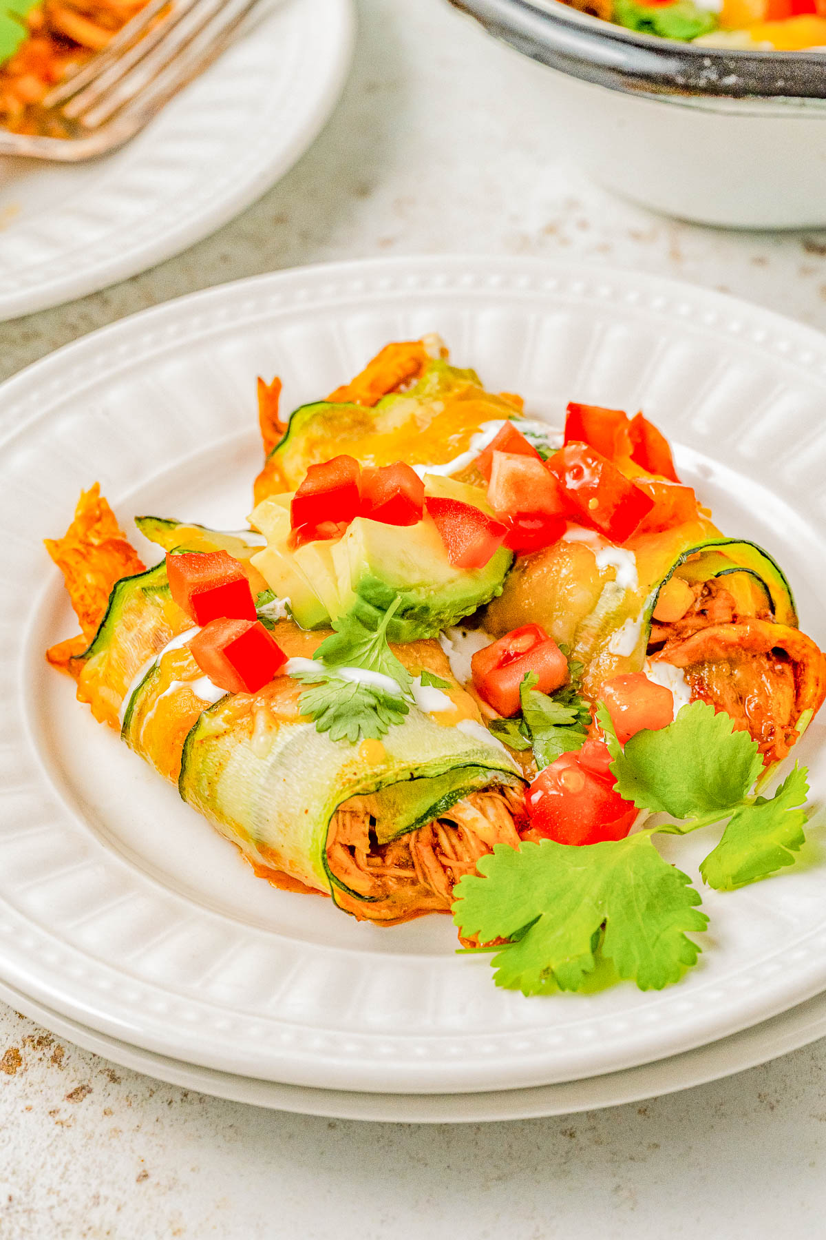 A white plate with zucchini enchiladas topped with melted cheese, avocado slices, chopped tomatoes, and cilantro.