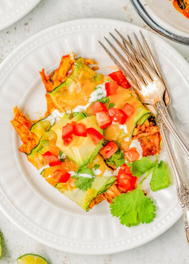 A white plate with zucchini enchiladas topped with melted cheese, diced tomatoes, avocado slices, sour cream, and cilantro. Two forks are placed on the side of the plate.