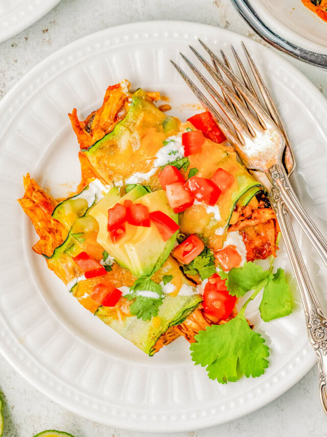 A white plate with zucchini enchiladas topped with melted cheese, diced tomatoes, avocado slices, sour cream, and cilantro. Two forks are placed on the side of the plate.