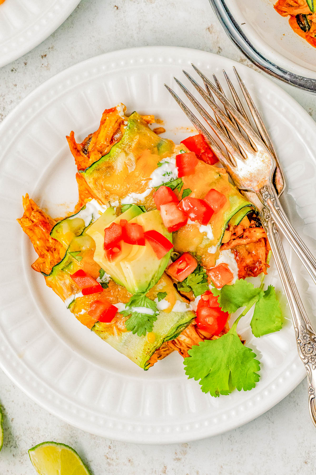 A white plate with zucchini enchiladas topped with melted cheese, diced tomatoes, avocado slices, sour cream, and cilantro. Two forks are placed on the side of the plate.