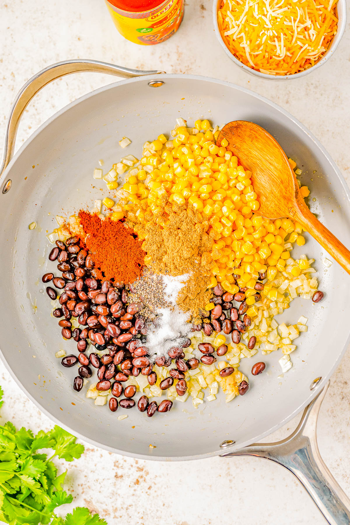 A pan contains black beans, corn, onion, and spices, with a wooden spoon. Surrounding the pan are a grated cheese bowl, an open jar of seasoning, and some cilantro leaves.