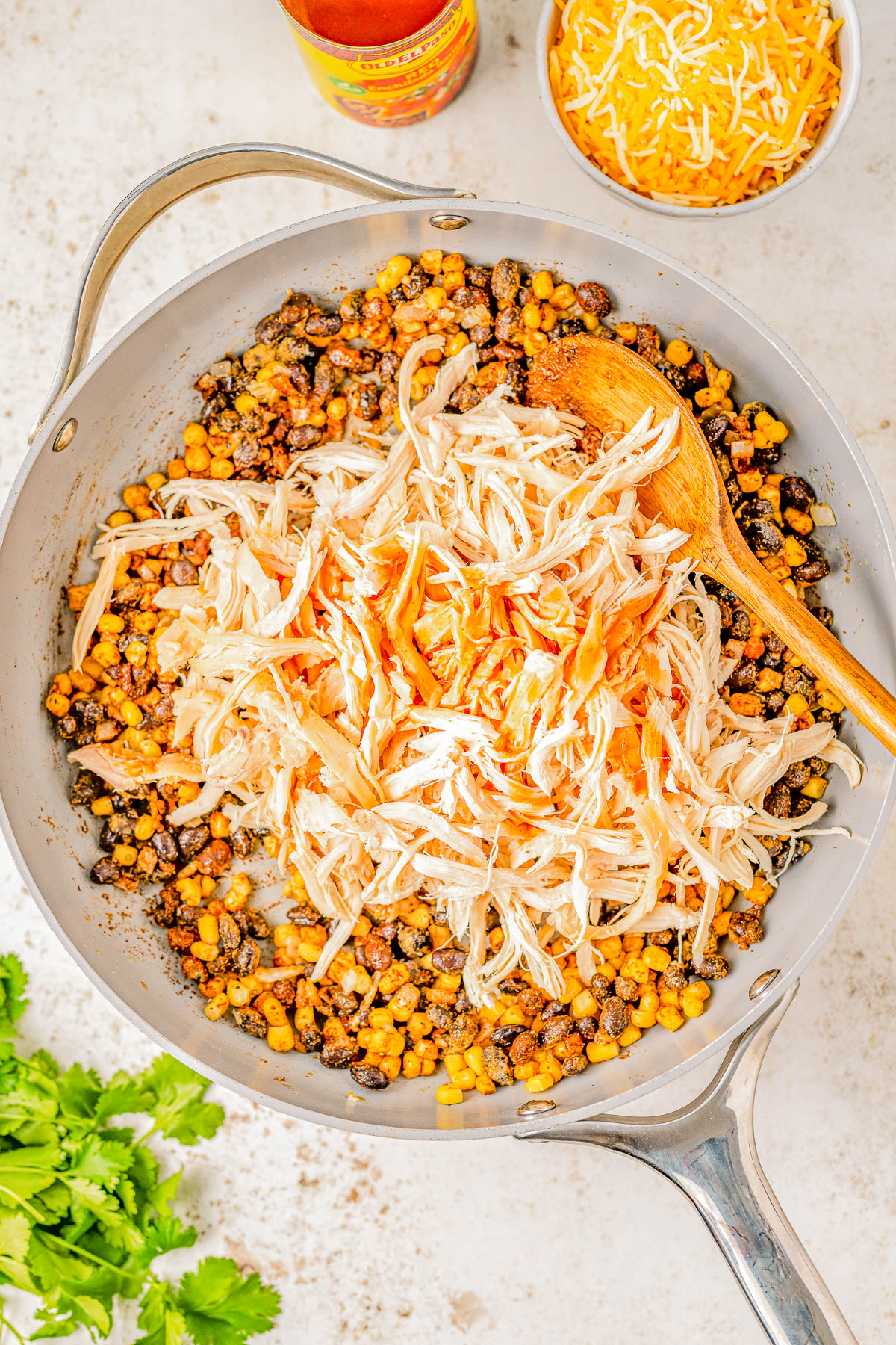 A skillet containing shredded chicken, corn, black beans, and hot sauce being stirred with a wooden spoon. Fresh cilantro and a bowl of shredded cheese are nearby.