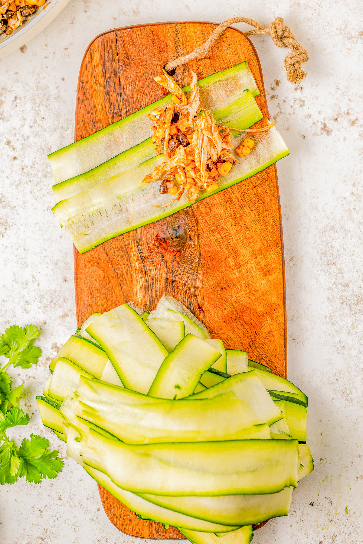 Thin slices of zucchini on a wooden cutting board with a small pile of nuts and dried tomatoes on top. A bunch of cilantro is partially visible to the side.