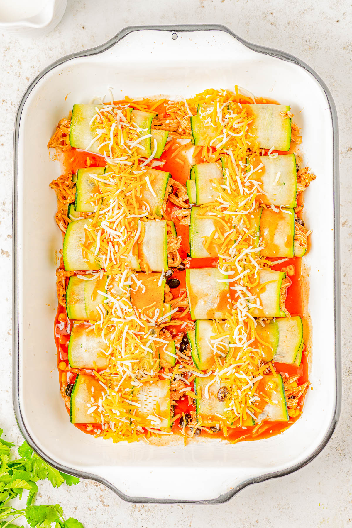 A white casserole dish filled with neatly arranged zucchini rolls, topped with shredded cheese and sauce, ready to be baked.