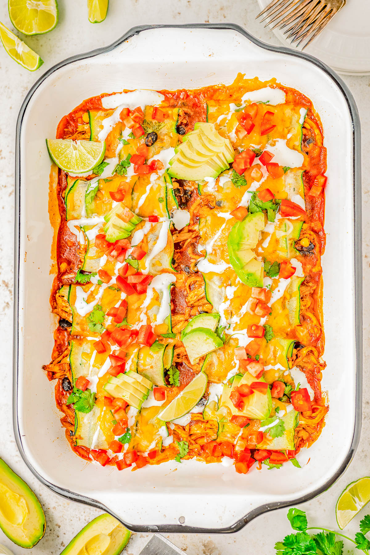 A casserole dish filled with enchiladas topped with melted cheese, diced tomatoes, avocado slices, lime wedges, and cilantro.