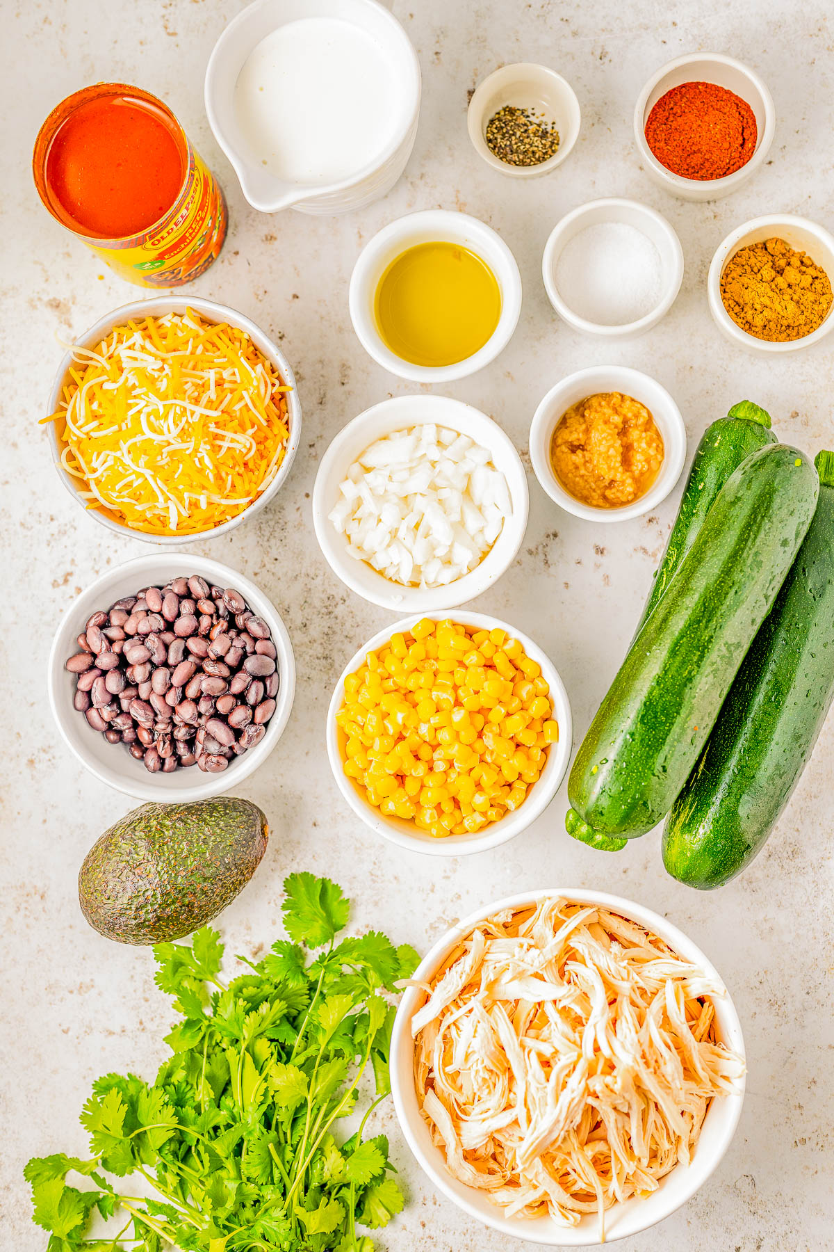 A flat lay of various ingredients including shredded chicken, zucchini, corn, black beans, grated cheese, avocado, cilantro, chopped onions, garlic, spices, olive oil, cream, and a can of tomatoes.