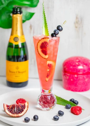 A tall cocktail garnished with blood orange slices and blueberries on a skewer, a champagne bottle in the background, and fruit pieces on a white plate.