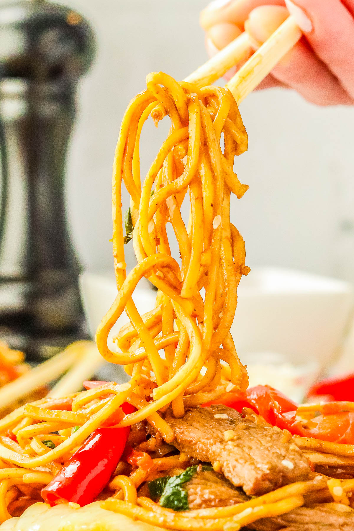 A hand using chopsticks to lift a portion of stir-fried noodles with vegetables and meat.