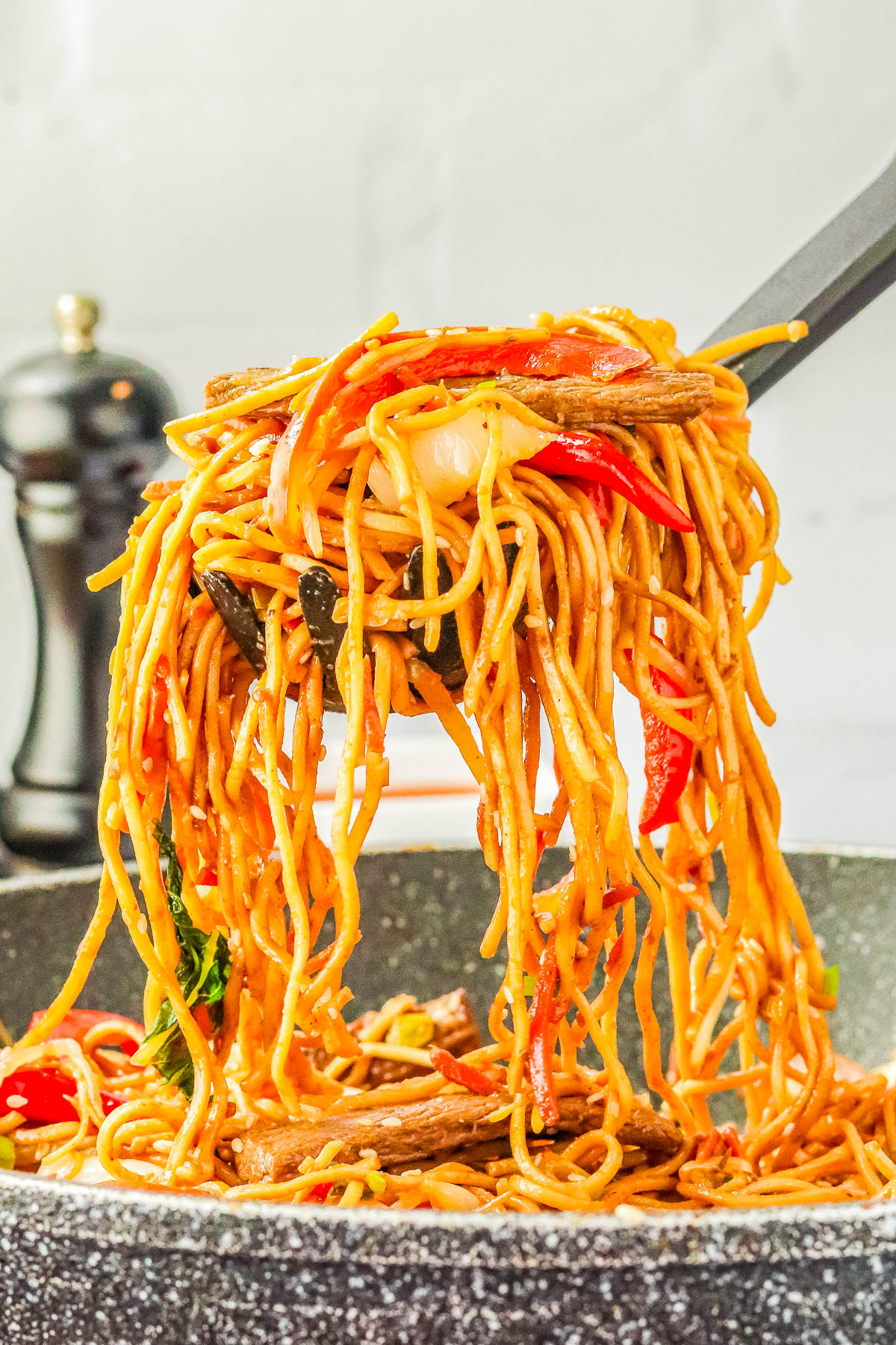 Close-up of a fork lifting a portion of vegetable stir-fried noodles from a speckled frying pan, with a pepper grinder in the background.