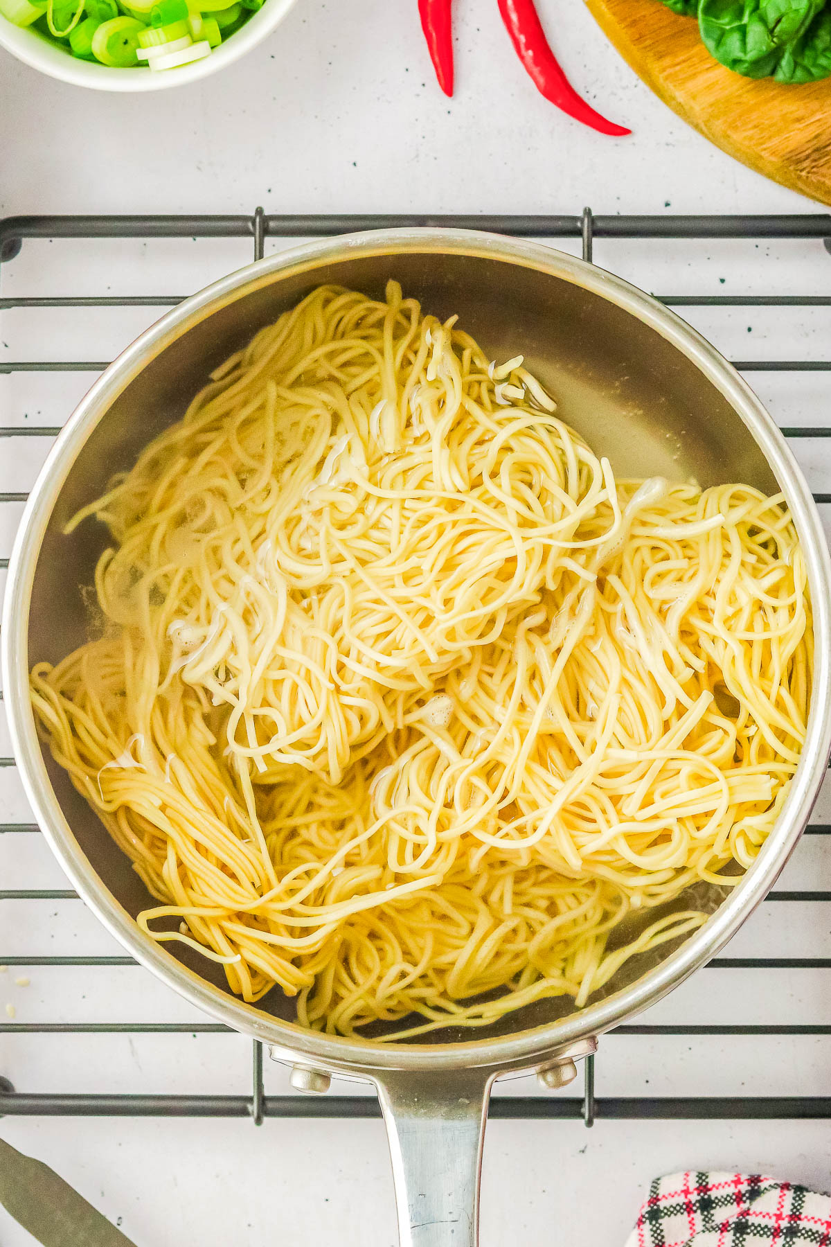 A pot containing freshly cooked noodles, seen from above on a cooling rack, with chopped green onions and chili peppers nearby.