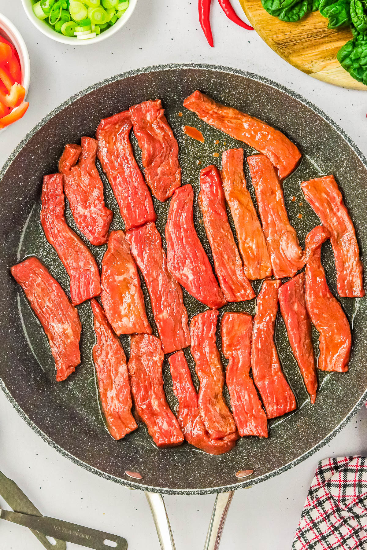 Sliced raw beef strips are arranged in a large frying pan. Various vegetables are visible in the background, including bell peppers, green onions, and spinach.