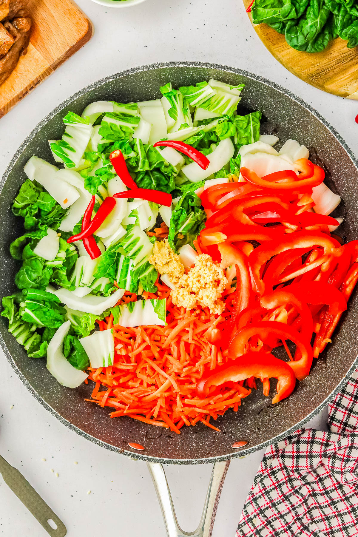 A pan contains a mix of chopped bok choy, red bell peppers, shredded carrots, and slices of red chili pepper, with minced garlic in the center. A checkered cloth is partially visible beside the pan.