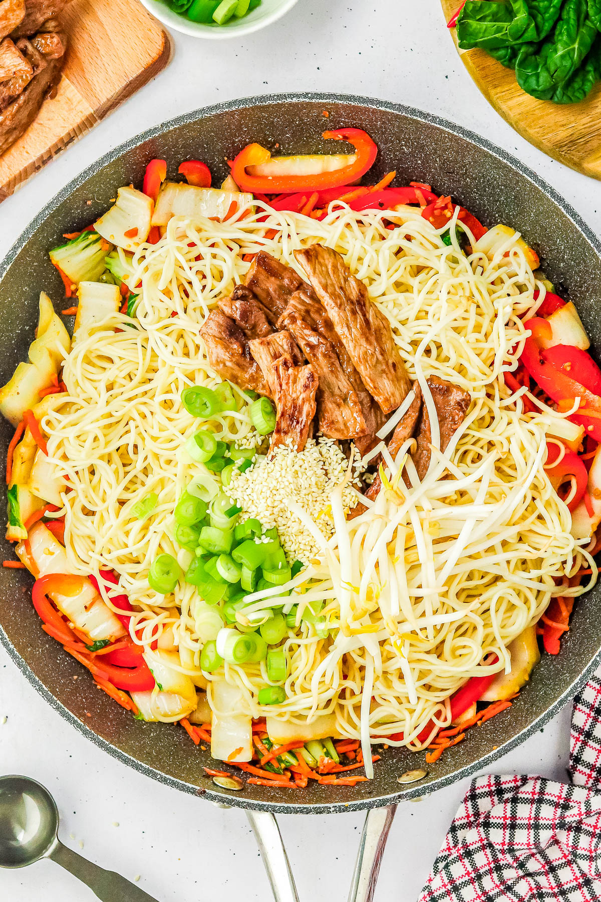 A skillet filled with cooked noodles, shredded vegetables, bean sprouts, green onions, sesame seeds, and pieces of meat, placed on a white surface with a napkin, spoon, and ingredients around.