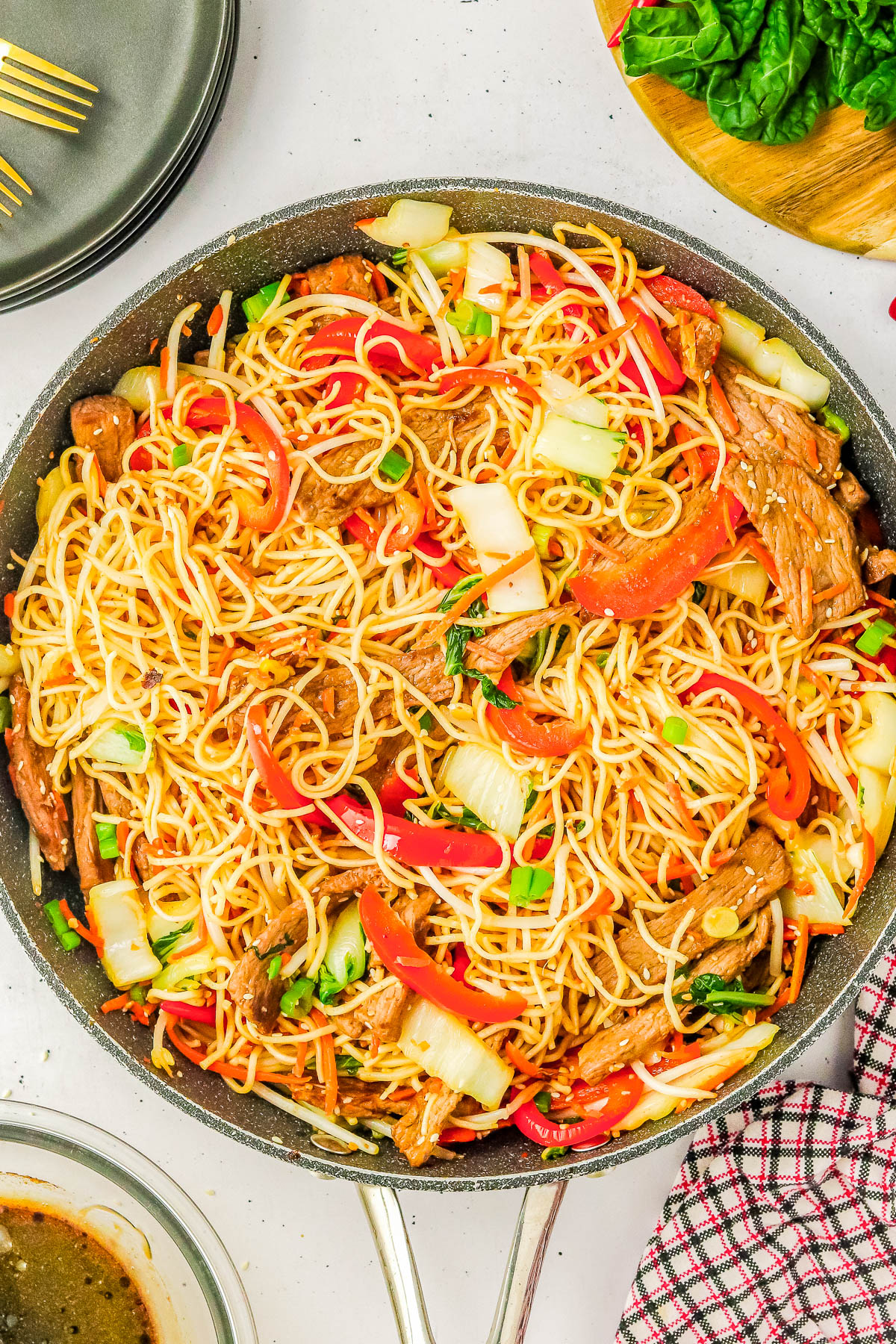 A pan filled with stir-fried noodles, sliced red peppers, bok choy, and beef strips. Plates, utensils, and fresh greens are visible around the pan.