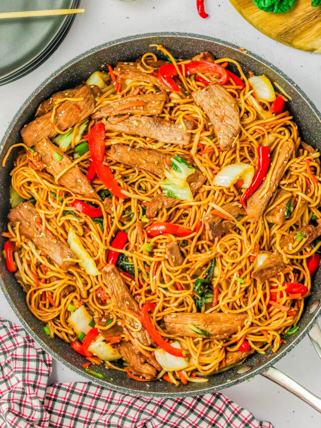 A pan of stir-fried noodles with slices of beef, red bell peppers, bok choy, and green onions. A red checkered cloth is underneath the pan. Plates and ingredients are visible in the background.