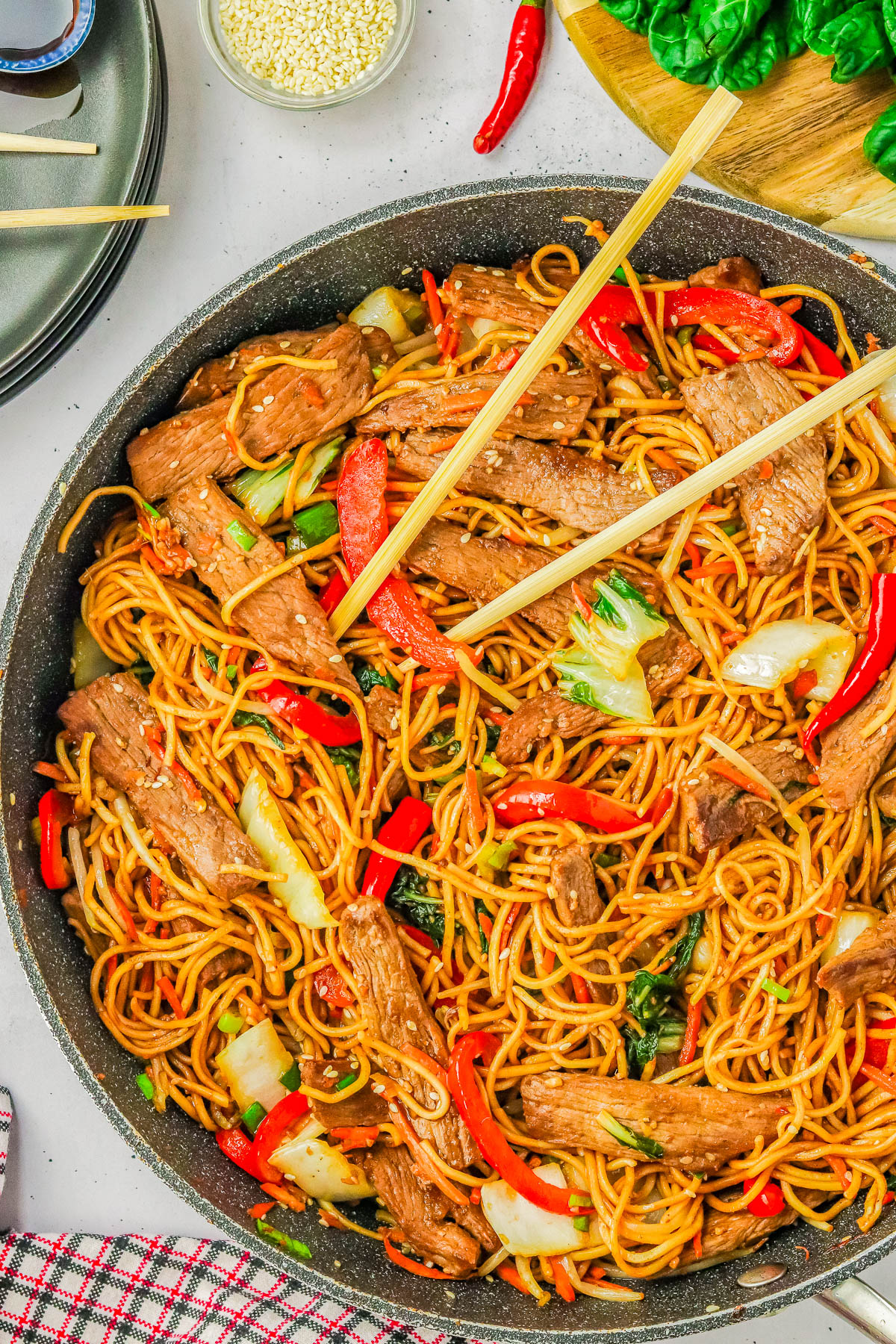 A pan of beef stir-fry with noodles, red bell peppers, cabbage, and garnished with sesame seeds. Chopsticks rest on top of the stir-fry. A small bowl of sesame seeds and leafy greens are visible nearby.